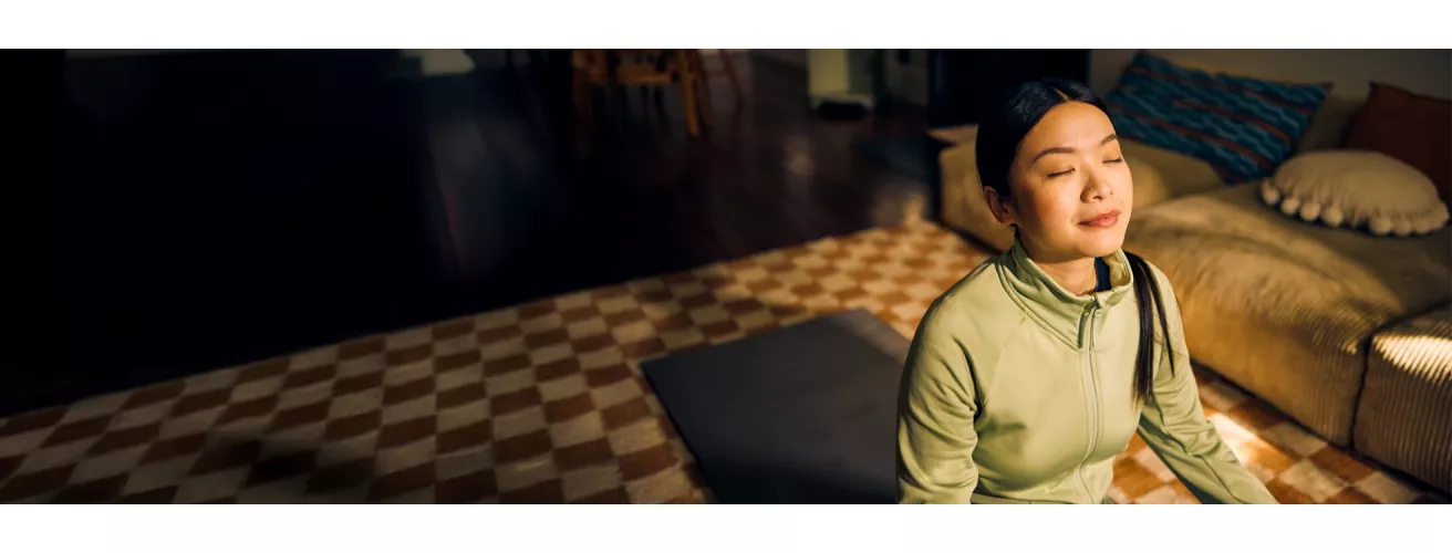 A person is seated on a yoga mat in a cozy indoor setting, wearing a light green top. The room features a checkered carpet, a beige sofa with cushions, and warm ambient lighting. The scene conveys a calm and serene atmosphere.