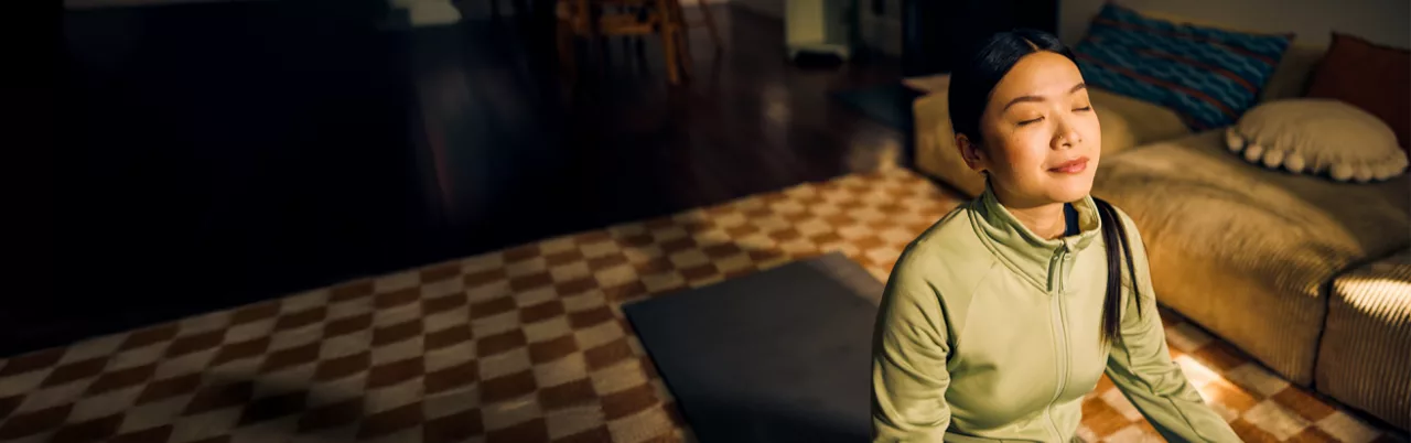 A person is seated on a yoga mat in a cozy indoor setting, wearing a light green top. The room features a checkered carpet, a beige sofa with cushions, and warm ambient lighting. The scene conveys a calm and serene atmosphere.