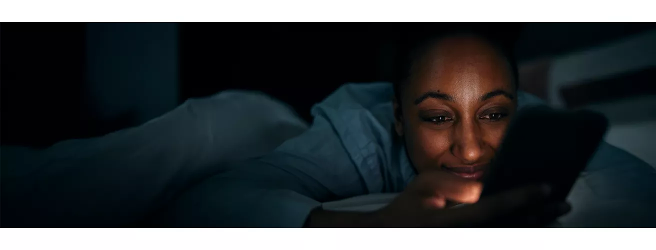 A person lies in bed at night, illuminated by the glow of a smartphone screen. The setting is a dimly lit bedroom, creating a quiet and intimate atmosphere. The focus is on the smartphone and the interaction, emphasizing late-night usage and technology in personal spaces.