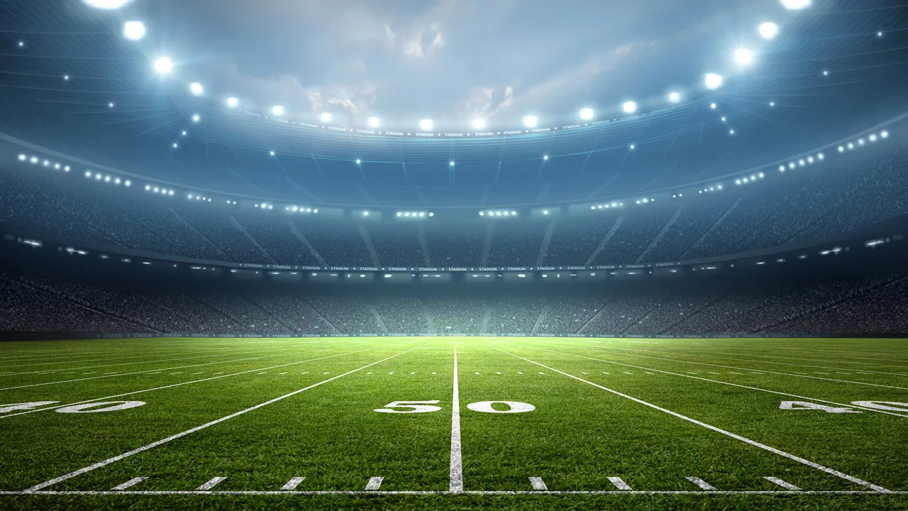 A brightly lit American football field is shown under powerful stadium lights at night. Yard lines and numbers are clearly visible on the green turf, including the 40, 50, and 60 yard markers. The stadium seating is silhouetted in the background, emphasizing the vast, empty arena. The overall scene conveys anticipation and energy before a big game.