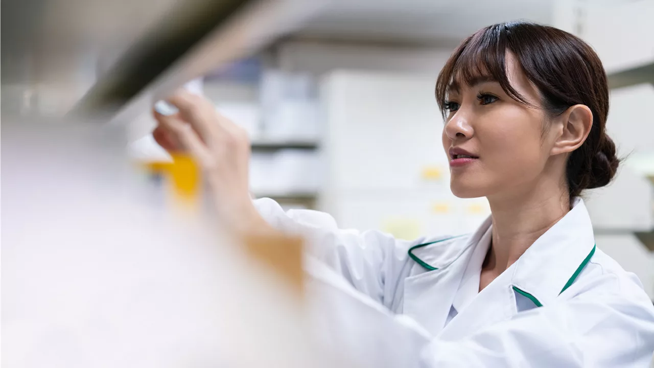 Woman in a lab coat reaching for items on a shelf in a pharmacy.