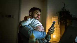 Father holding a sleeping baby while using his phone late at night