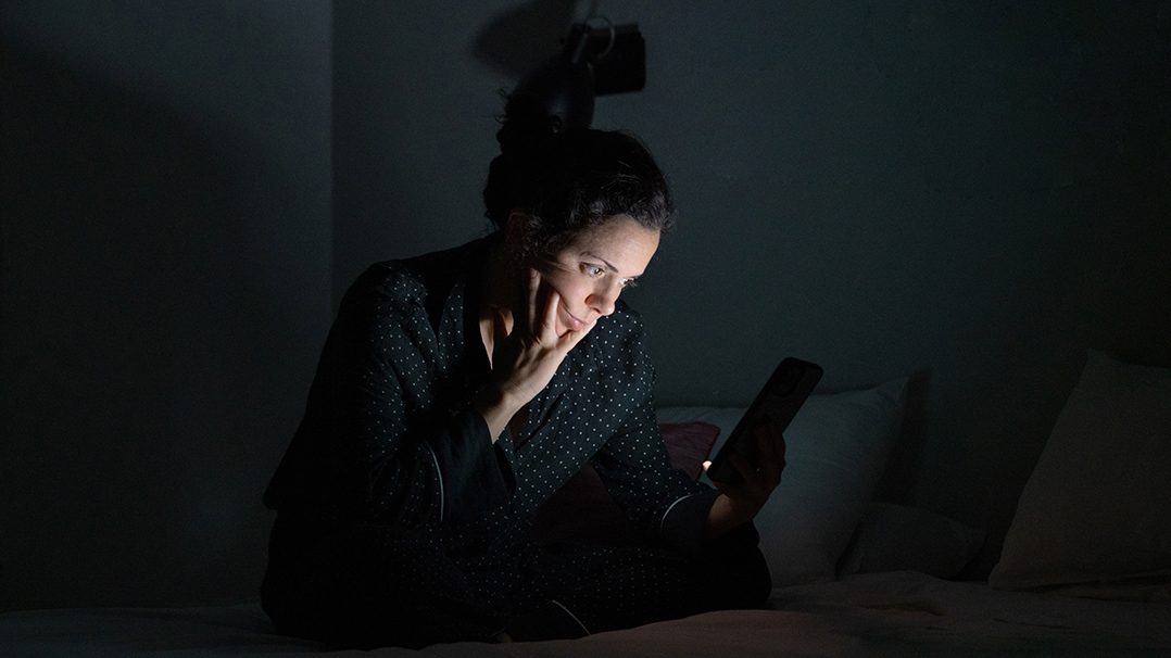 A person sits on a bed in a dimly lit room, holding a smartphone. The setting suggests nighttime or low-light conditions, with the glow of the phone screen illuminating their hands. The individual appears to be wearing dark, casual sleepwear, and the background includes pillows and minimal decor.
