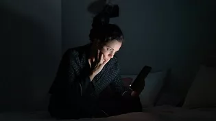 A person sits on a bed in a dimly lit room, holding a smartphone. The setting suggests nighttime or low-light conditions, with the glow of the phone screen illuminating their hands. The individual appears to be wearing dark, casual sleepwear, and the background includes pillows and minimal decor.