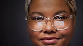 A person wearing thin-framed glasses is partially visible against a dark background. The image highlights the glasses and a portion of the person's hair. The setting appears minimalistic with no additional visible elements or text.