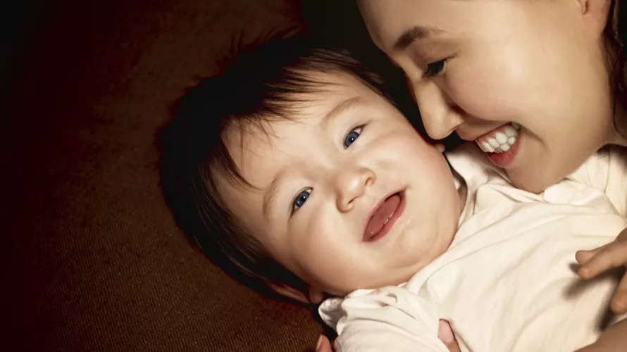 A baby wearing a plain white onesie is lying on a soft brown surface. The setting appears warm and cozy, with a focus on the infant's clothing and the textured background. The image conveys a sense of comfort and simplicity.