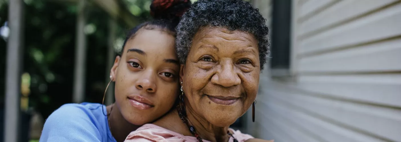 Two individuals are seen embracing outdoors near a residential building with visible siding. The setting includes greenery in the background, suggesting a suburban or natural environment. The image conveys warmth and connection, with casual clothing and soft lighting enhancing the mood.