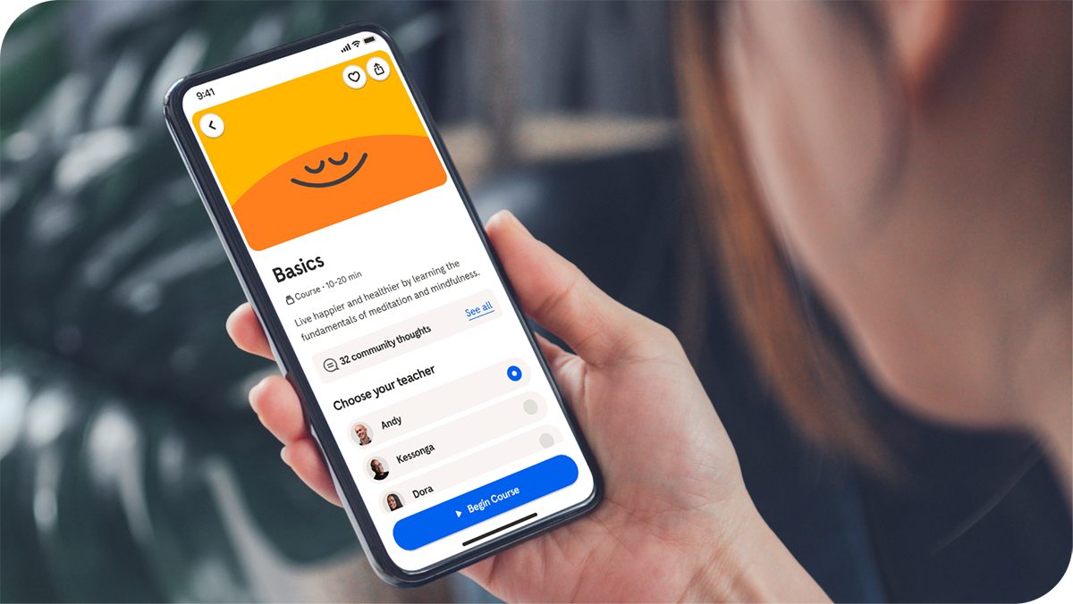 A person holds a smartphone displaying a meditation or mindfulness app with the title 'Basics' and an orange graphic. The screen shows options to choose a teacher and a visible blue 'Start Course' button. The setting appears to be indoors with a blurred background, and the time on the phone reads 9:41.