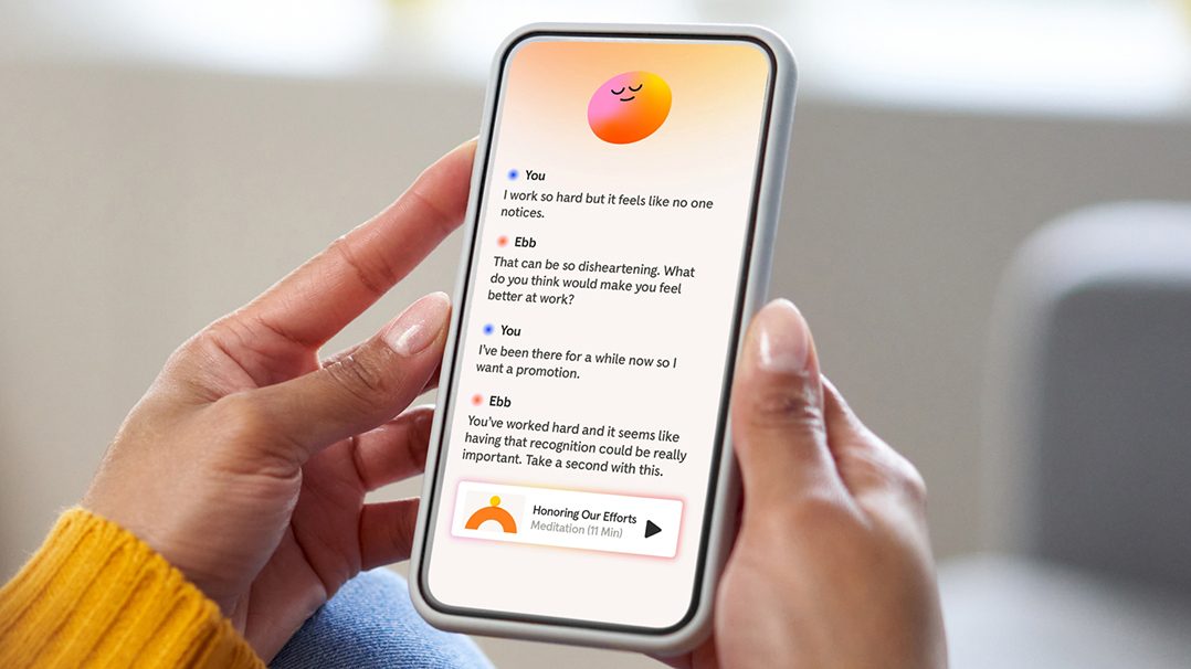 A person holds a smartphone displaying a mental health or wellness app with a chat interface. The screen shows a conversation about work recognition and a suggestion to honor efforts, with a visible button labeled 'Honoring Our Efforts Meditation 10 Min'. The individual is seated indoors, wearing a yellow sweater, and the setting appears relaxed and casual.