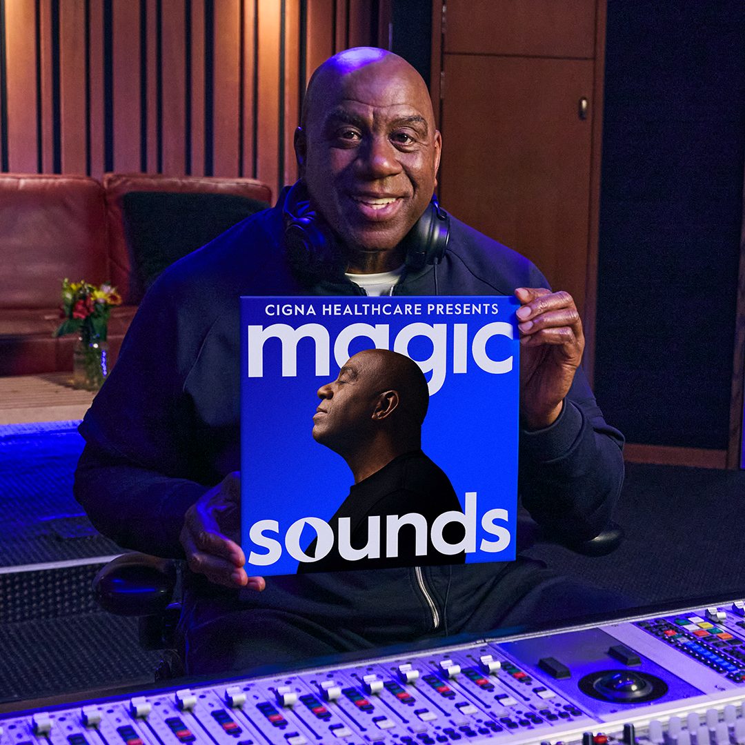 Magic Johnson is seated in a professional recording studio holding a poster titled 'Magic Sounds' presented by Cigna Healthcare. The studio features a mixing console and wooden paneling, creating a creative and professional atmosphere. The poster displays bold text and a silhouette of a person against a blue background.