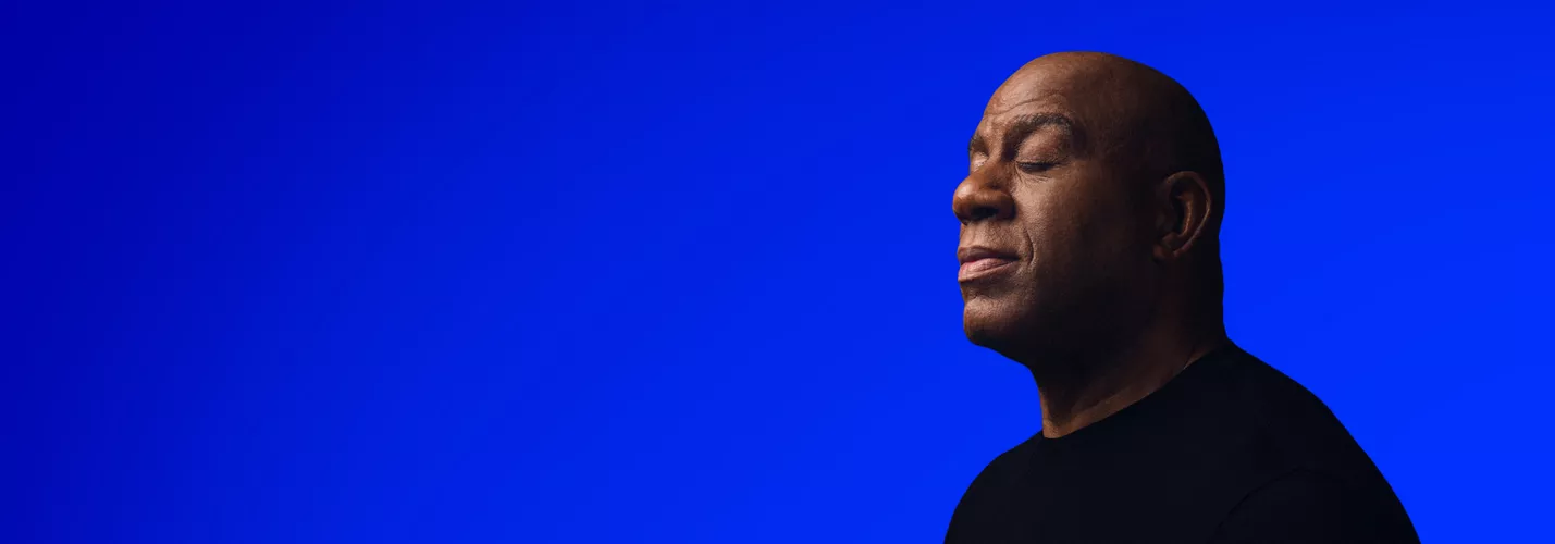 Magic Johnson wearing a black shirt is positioned against a vibrant blue backdrop. The image features a minimalist and modern aesthetic with strong color contrast. The setting emphasizes simplicity and bold visual appeal.