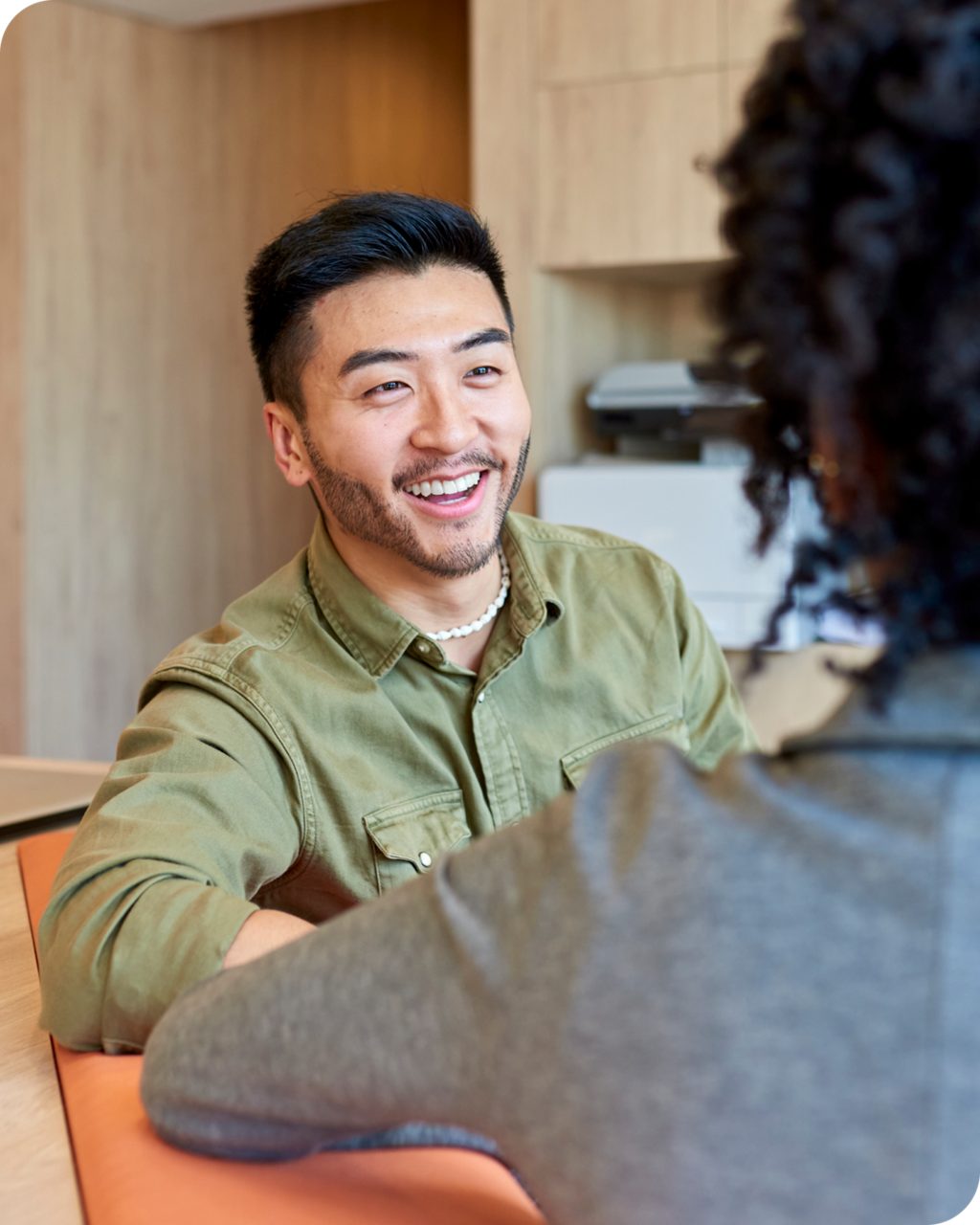 Two individuals are engaged in a conversation in a contemporary office environment. The setting features wooden furniture and a printer in the background, suggesting a professional workspace. One person wears a green button-up shirt, while the other is dressed in a gray outfit. The image conveys a casual yet professional mood.