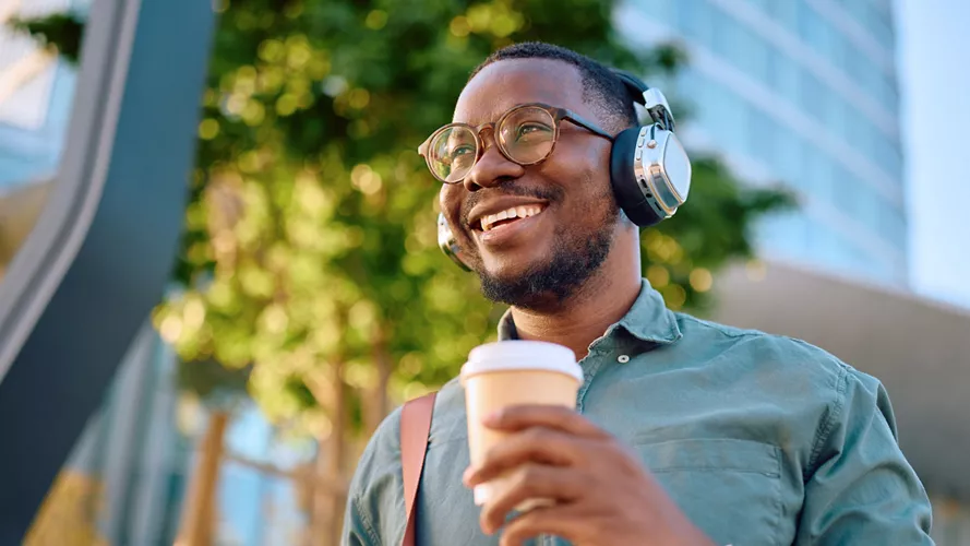 A person is seen outdoors wearing large over-ear headphones and holding a takeaway coffee cup. The setting appears to be urban with greenery and modern buildings in the background, suggesting a city environment. The individual is dressed casually in a button-up shirt and has a shoulder strap visible, possibly from a bag. The lighting is bright and natural, evoking a relaxed and energetic mood.