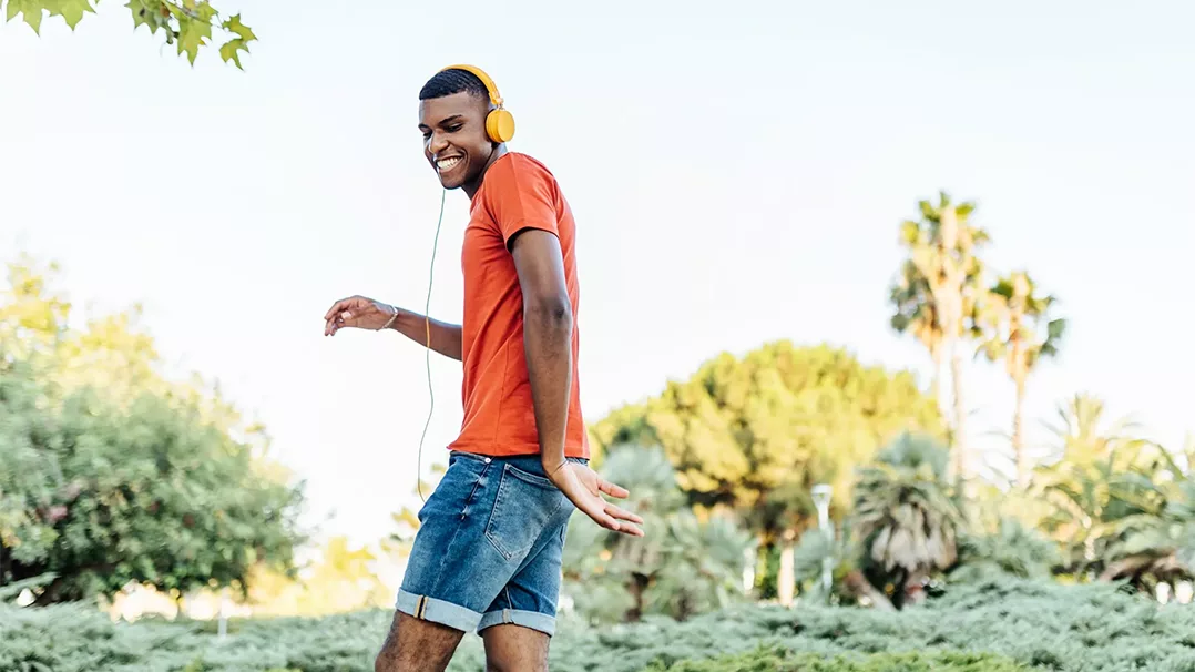 Young man in an orange shirt and yellow headphones dances joyfully outdoors.