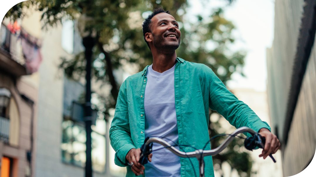 A casually dressed man walks a bicycle along an urban street. He wears a green shirt over a white t-shirt while holding the handlebars of the bike. The background shows buildings, trees, and a slightly blurred streetscape, suggesting motion and a city environment. The overall scene feels relaxed and contemporary, with natural daylight and soft focus on the surroundings.