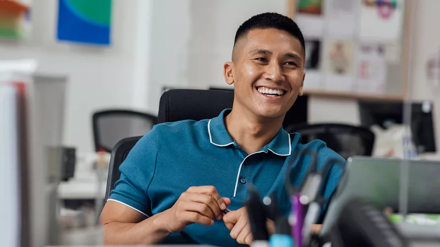 A man wearing a teal polo shirt with white trim is seated at an office desk. The setting appears to be a modern workspace with blurred background elements including colorful wall art and office supplies. The desk features pens, a laptop, and other items, suggesting a professional environment.