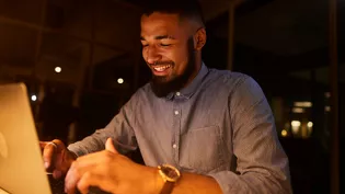 A man wearing a blue button-up shirt is focused on working on a laptop in a dimly lit indoor setting. The warm lighting creates a cozy and professional atmosphere. A gold wristwatch is visible on his left wrist, adding a touch of sophistication.