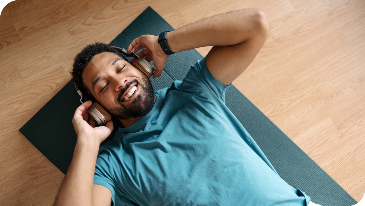 A man is lying on a dark green yoga mat listening to music in a bright indoor setting. He is wearing a teal t-shirt and a smartwatch on his wrist. The wooden floor adds a warm tone to the environment, emphasizing a casual atmosphere.