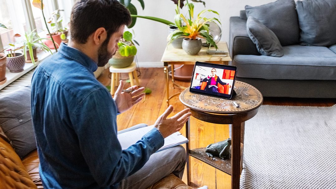 A man sits in a well-lit living room, engaging in a video call with a therapist on a tablet.