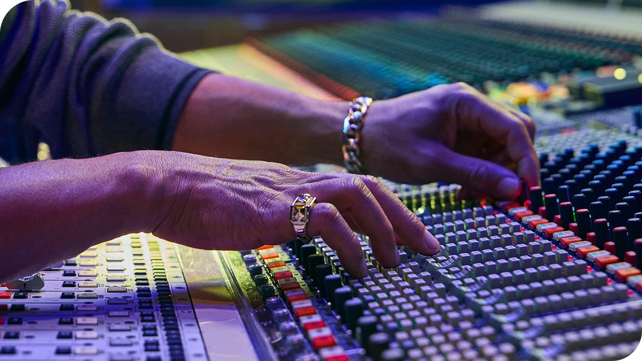 Close-up of hands operating a professional audio mixing console in a studio setting. The scene features vibrant lighting and detailed controls, emphasizing precision and technical expertise. The individual wears a bracelet and ring, adding a personal touch to the composition.