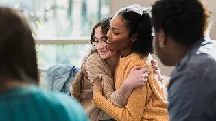 Two women hugging as part of a small group. They are both wearing sweaters and have their eyes closed, with slight smiles on their faces, indicating comfort.
