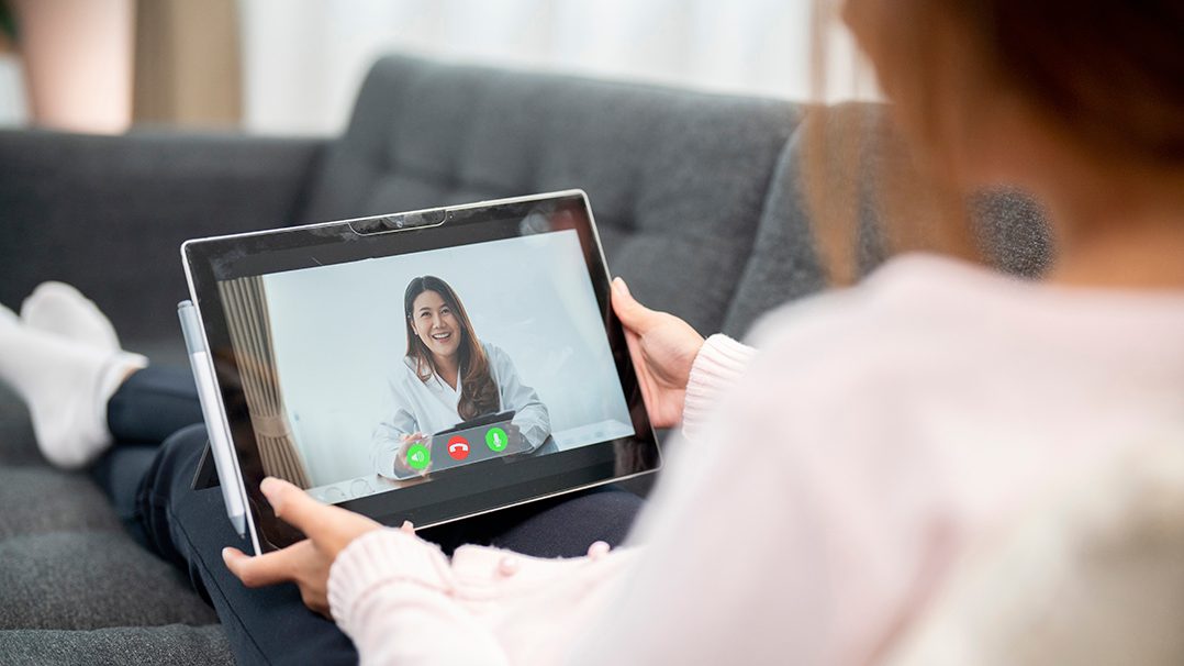 A woman is seated on a couch holding a tablet device during a video call. The screen displays a person in a white coat, suggesting a virtual consultation or meeting. The setting is a cozy living room with soft lighting and casual attire.
