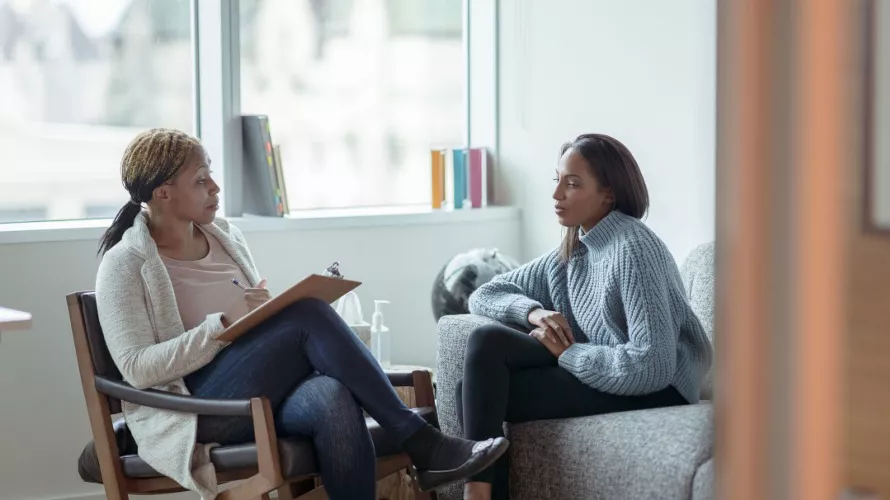 Two women are seated in a professional office environment, engaging in a counseling session. One woman holds a clipboard, suggesting note-taking or assessment, while the other listens attentively. The setting includes a cozy chair, a sofa, and a window with books and decor visible. A bottle of hand sanitizer is placed on the table, emphasizing hygiene.