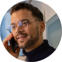 A man holds a smartphone to his ear while engaged in a phone call. He is wearing a dark turtleneck sweater and glasses, with his face partially obscured. The background shows an indoor setting with soft, neutral colors and a contemporary feel. The close-up composition emphasizes communication and concentration.
