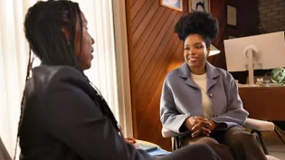 Two women sit facing each other in a warm, wood-paneled office setting. One woman listens attentively while the other speaks, suggesting a counseling or professional conversation. The room includes a desk with a computer monitor and decorative items in the background. Soft lighting and neutral tones create a calm, comfortable atmosphere.