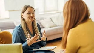 Two women sit facing each other in a bright living room during a counseling session. The woman in blue gestures with her hands while holding a pen and notebook, suggesting active listening and guidance. The other woman, with long red hair, listens attentively from a nearby chair. Soft furnishings and natural light create a calm, comfortable atmosphere suitable for therapy or coaching.