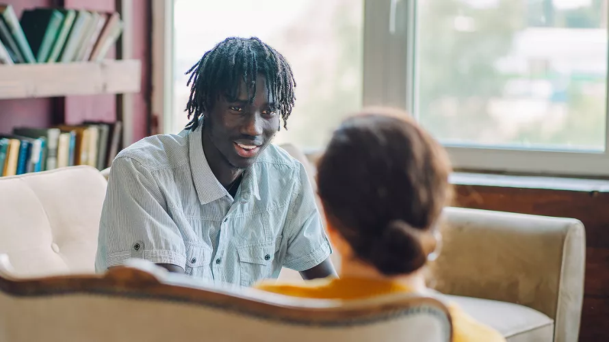 Two individuals are seated in a comfortable room with bookshelves and a large window in the background. The setting suggests a counseling or therapy session, with one person attentively listening to the other. 