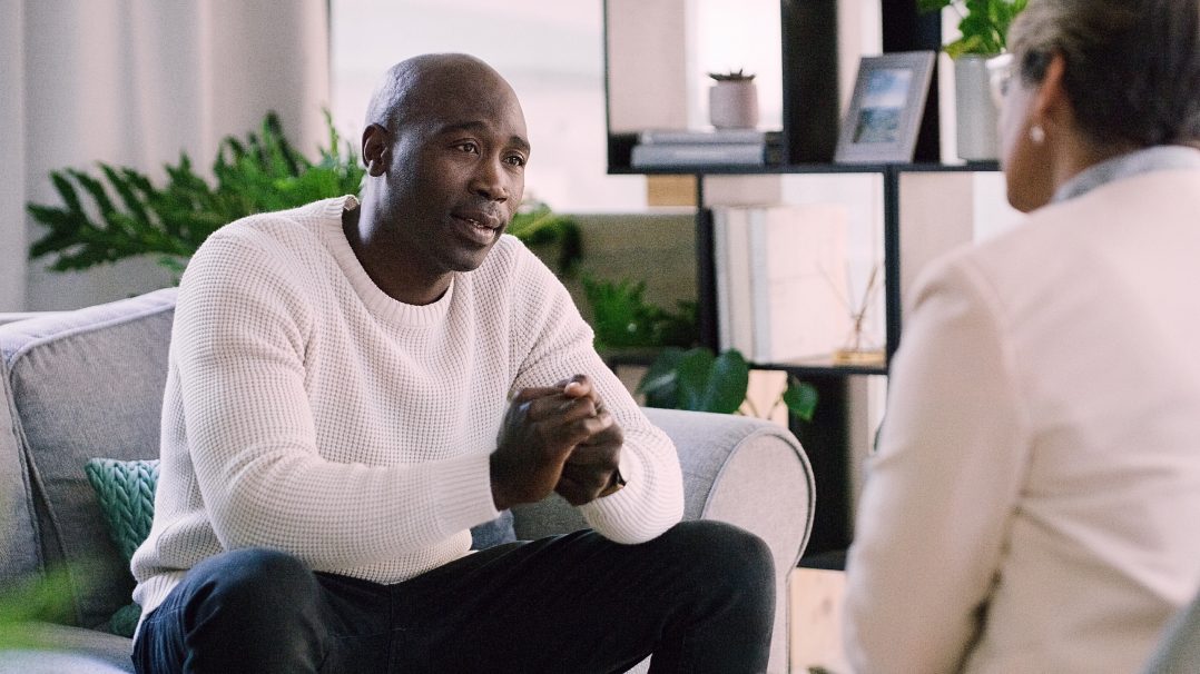 A man in a white sweater sits on a gray couch, engaged in conversation with another person in a light-colored blazer. The setting appears to be a modern, well-lit living room or office space with indoor plants and shelves in the background. The atmosphere suggests a professional or therapeutic interaction.