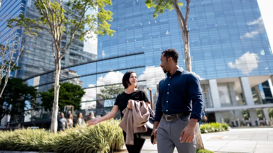 Two individuals are walking and conversing in an urban setting with a modern glass building in the background.