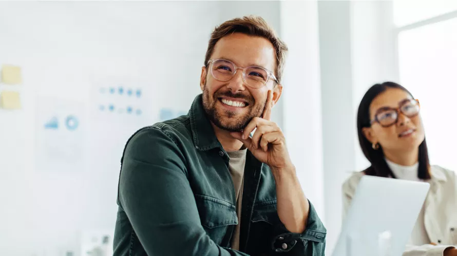 A professional setting featuring two individuals in a modern office environment. The focus is on a man in a green jacket seated at a table with a laptop and documents. The background includes bright natural light and blurred charts on the wall, suggesting a collaborative workspace.