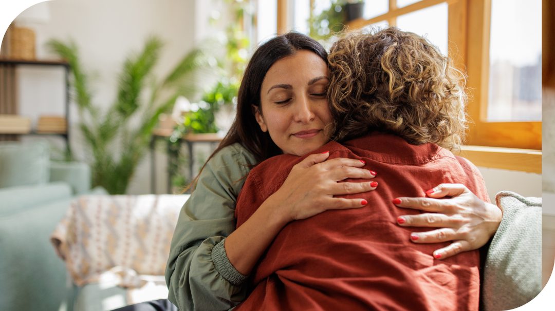 Two people sit closely together on a couch, embracing in a warm hug. The scene takes place in a bright living room with large windows and natural daylight streaming in. Green plants and soft furnishings create a cozy, relaxed atmosphere. The image conveys comfort, support, and emotional connection between the individuals.