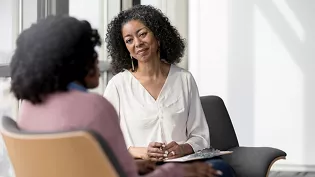 Two individuals are engaged in a counseling or discussion session in a bright, modern office space. The setting features natural light streaming through large windows and contemporary furniture. The focus is on the interaction, with one person holding a clipboard, suggesting a professional or therapeutic context.