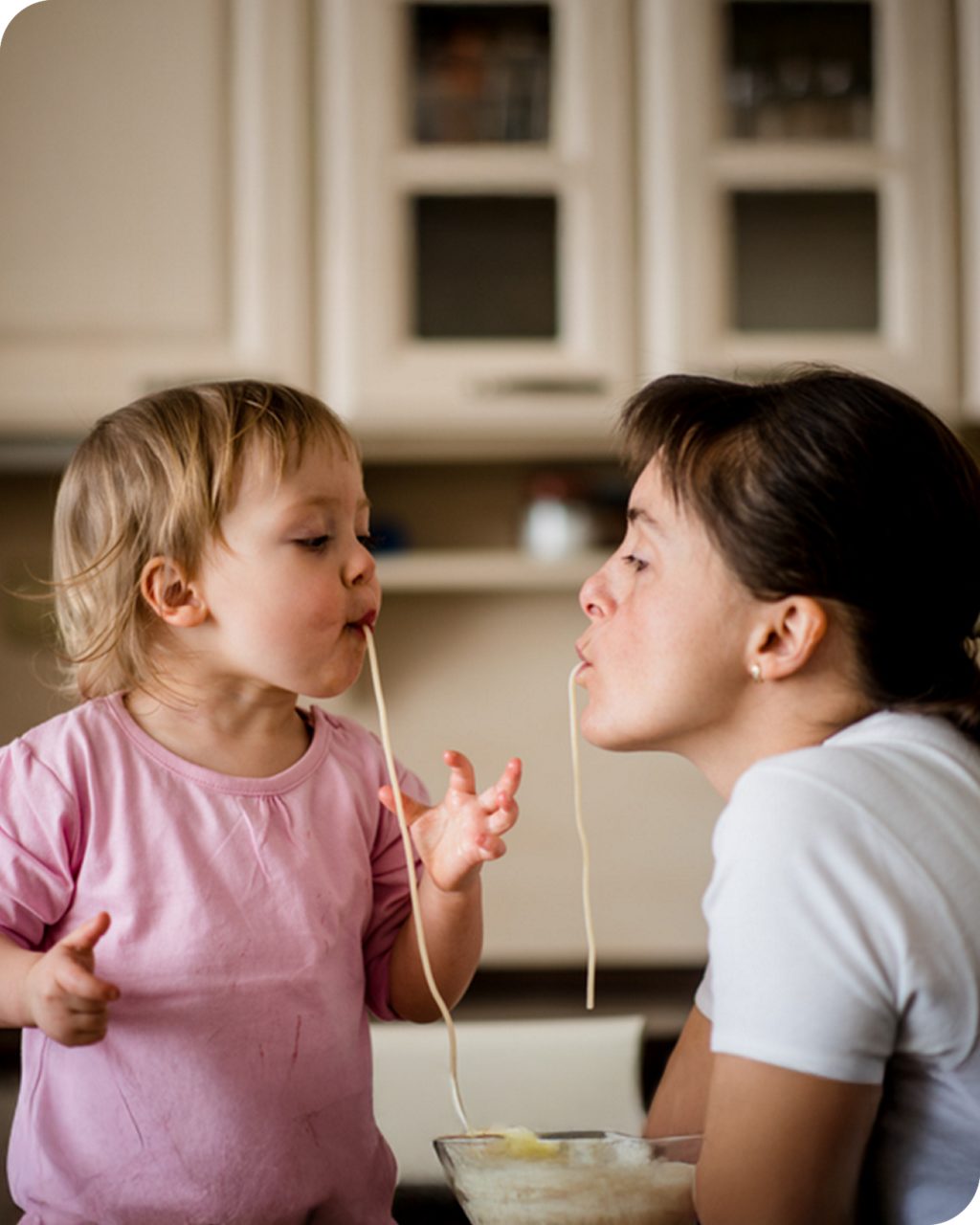 A young child in a pink shirt and an adult in a white shirt are sharing a meal of spaghetti in a cozy kitchen setting. The child is holding strands of spaghetti while engaging with the adult. The background features light-colored cabinets, adding a warm and homely atmosphere.