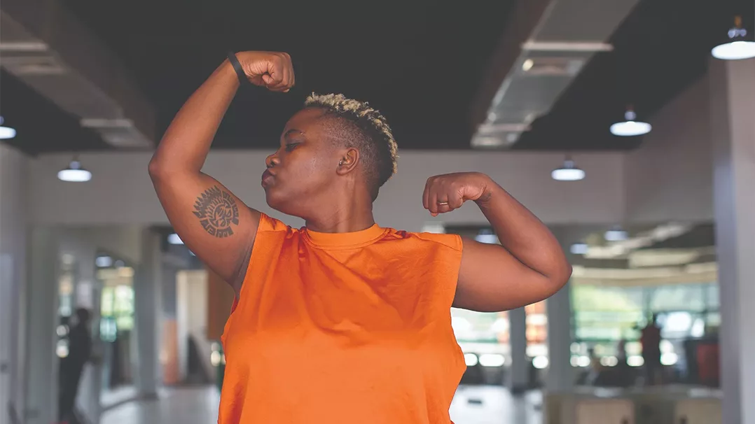 A person in an orange shirt flexes their arms in a gym setting.