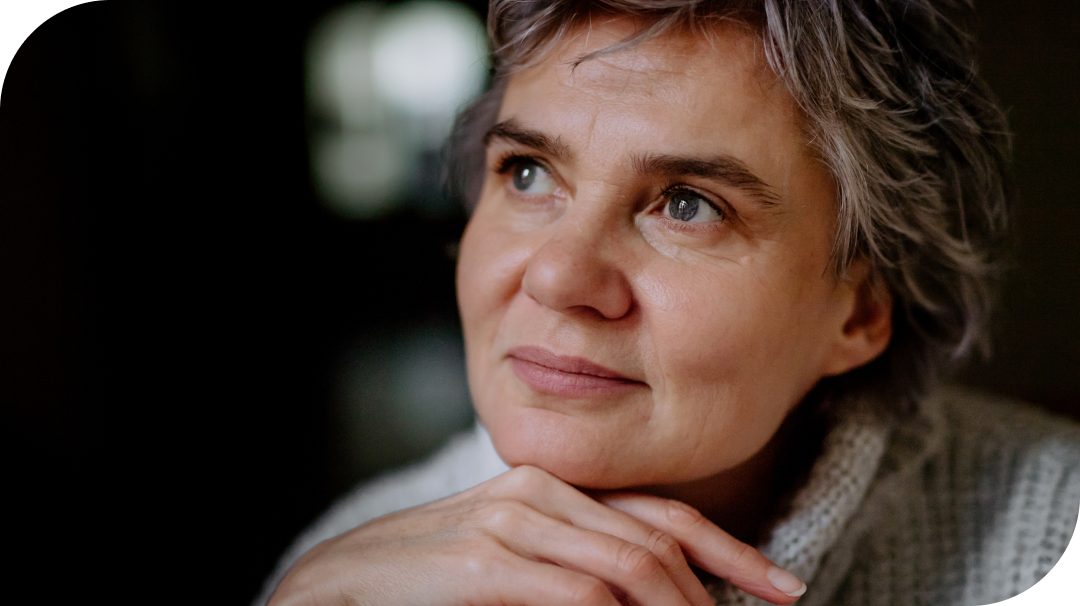 A person with short, gray hair rests their hand near their face in a contemplative pose. The background is dark and softly blurred, drawing attention to the subject. Soft, natural light highlights the texture of their sweater and hair. The overall scene feels quiet and introspective.