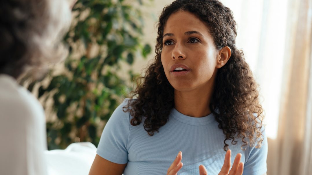 A woman wearing a light blue shirt is seen gesturing with her hands during a conversation. The setting appears to be indoors with natural light and a blurred background featuring a plant. The scene conveys a casual and engaging atmosphere.