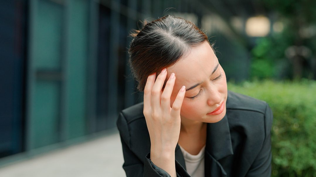 A woman dressed in a black blazer and white shirt is seated outdoors in an urban setting. The background features modern architecture and greenery, suggesting a professional environment. Her posture conveys a contemplative or stressed mood.