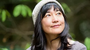 A woman wearing a beige knit hat and a gray sweater is pictured outdoors in a lush, green natural setting. The background features soft-focus foliage, creating a serene and tranquil atmosphere. The image emphasizes casual style and a connection to nature.