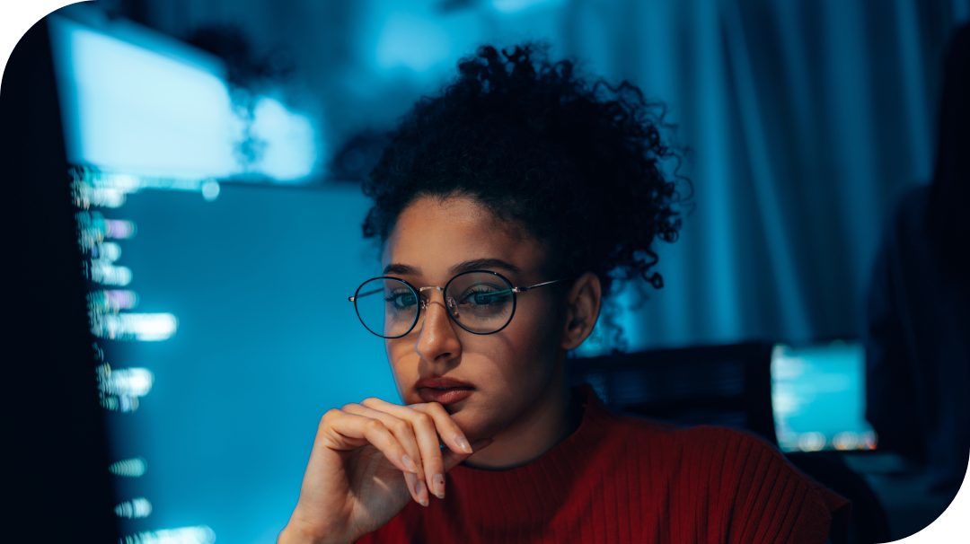 A person sits at a desk in a dimly lit office, looking at large computer monitors filled with code. The subject’s face is obscured, emphasizing the technology and work environment rather than identity. Cool blue light from the screens contrasts with the warm color of the person’s clothing, creating a focused, nighttime atmosphere. The scene suggests concentrated programming or cybersecurity work in a modern workspace.