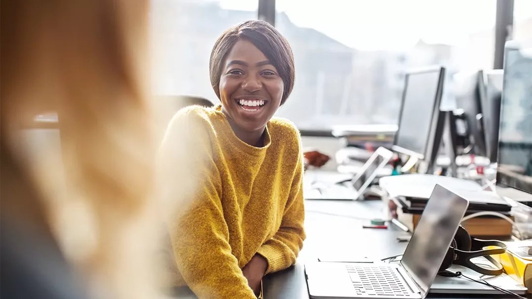 Smiling woman in a yellow sweater sitting at a desk with multiple computers.