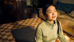 A woman wearing a light green zip-up jacket is seated on a yoga mat in a cozy indoor setting. The room features warm lighting, a checkered rug, and a beige couch with striped cushions in the background. The scene suggests a calm and relaxed atmosphere, ideal for meditation or mindfulness activities.