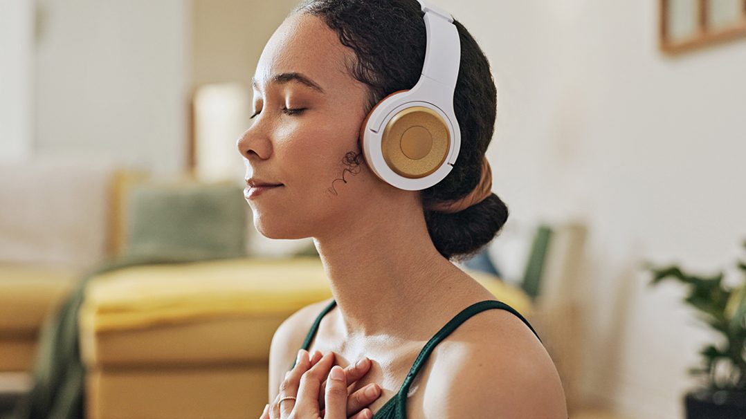 A woman is seated indoors, wearing large over-ear headphones with gold accents. She holds her hands to her chest in a gesture of calm or meditation, suggesting a mindfulness or relaxation activity. The background features a cozy living space with soft lighting and neutral tones, contributing to a peaceful atmosphere.