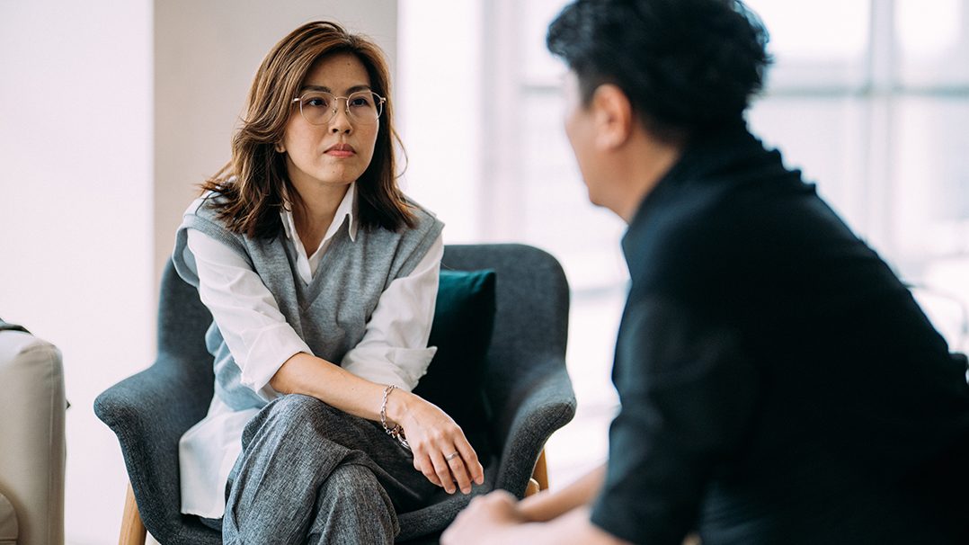 Two individuals are engaged in a professional conversation in a modern office environment. The woman is seated on a gray armchair, wearing a white shirt and gray vest, while the man is partially visible in the foreground. The setting features soft natural lighting and minimalistic decor, creating a calm and focused atmosphere.