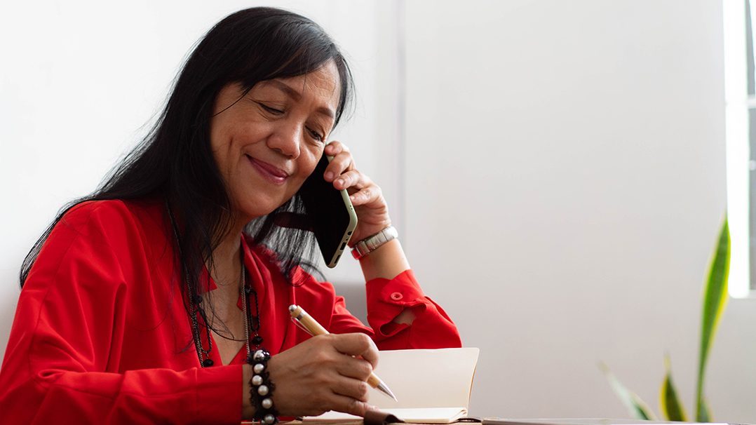 A woman wearing a vibrant red shirt is seated at a desk, holding a pen and writing in a notebook while speaking on a smartphone. The setting appears to be a bright indoor space with natural light and a minimalistic decor, including a green plant in the background. She is accessorized with bracelets and a necklace, adding a touch of personal style.