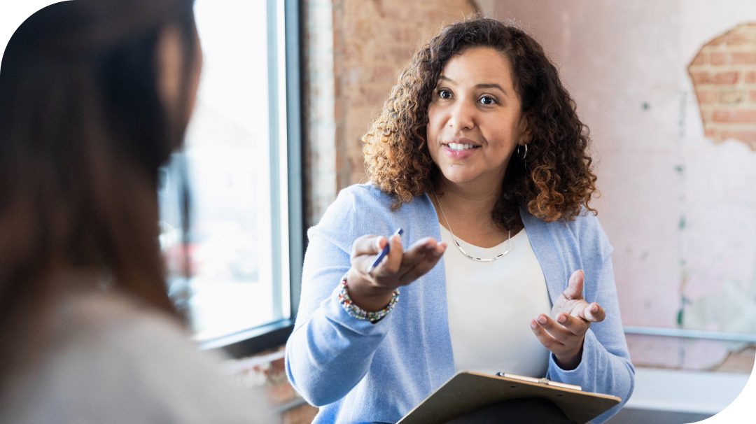 A professional woman sits indoors holding a clipboard and pen while speaking to another person. She appears to be explaining or clarifying something with an open hand gesture. The setting looks like an office or counseling space with exposed brick walls and soft natural light. The focus is on communication and support between the two individuals.