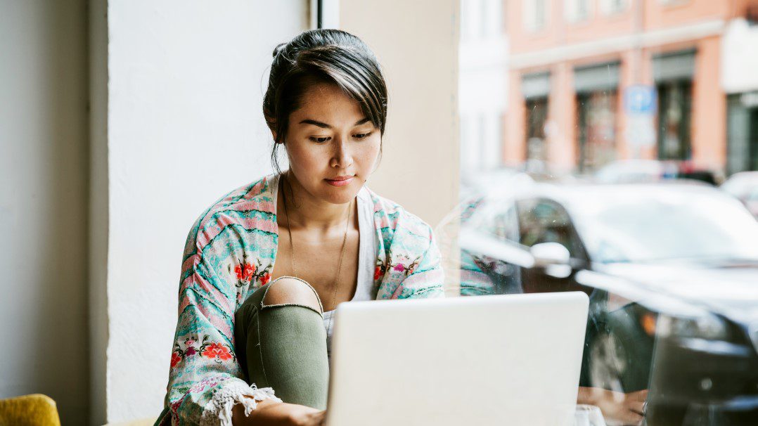 A young woman sits indoors near a large window, using a laptop in a casual setting. She is wearing a floral-patterned cardigan and green pants, with natural light streaming in. The background shows parked cars and urban architecture outside the window.